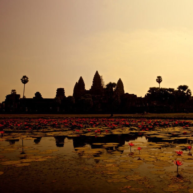 The ancient Cambodia temple silhouette is reflected in the pond with lotus flowers at sunset