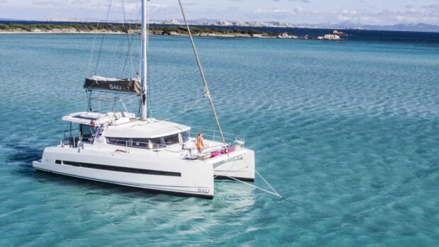 A catamaran sailing on clear blue water near a rocky coastline with a person on board