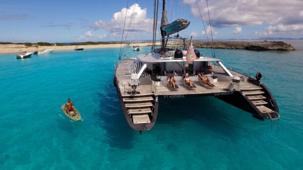 A catamaran floating on clear blue water with people relaxing and a man kayaking nearby