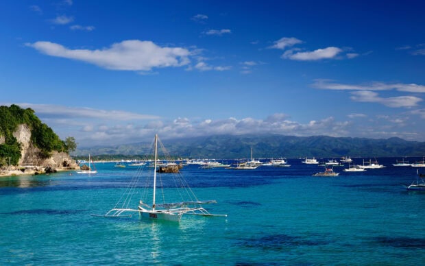 Catamaran sailing on clear blue water near a rocky coastline under a bright sky with distant mountains