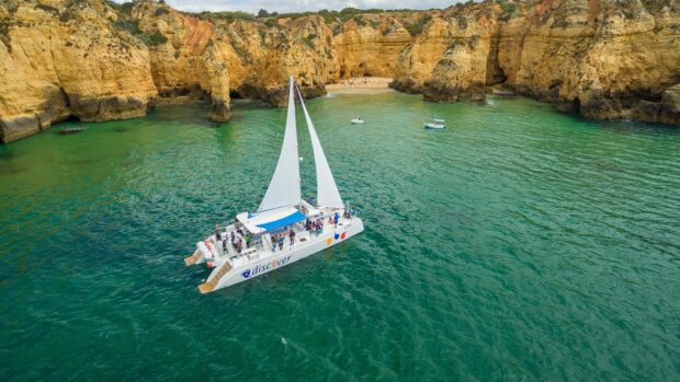 White catamaran sailing near rocky cliffs in clear green water with passengers on board
