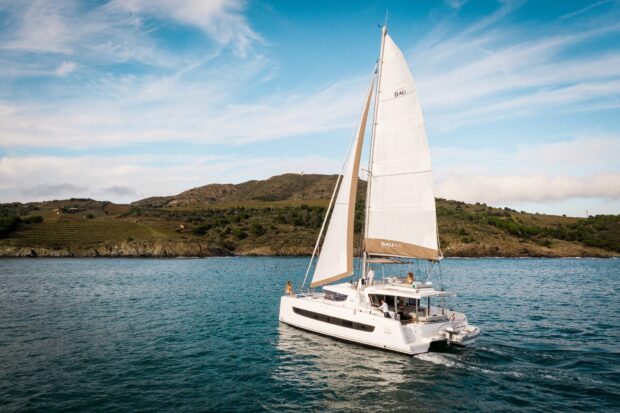White catamaran sailing on calm blue sea near a green hillside