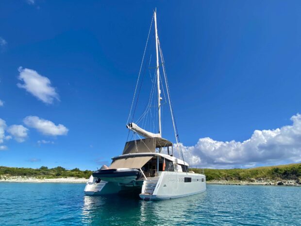 A catamaran anchored in clear blue water near a green shoreline under a bright blue sky