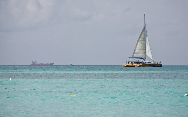 A catamaran sailing on clear turquoise water under a cloudy sky with distant ships in the background
