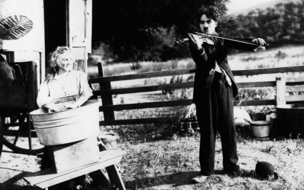 Charlie Chaplin plays violin outdoors near a woman washing clothes in a tub