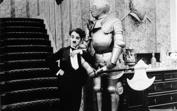 Charlie Chaplin stands in a tuxedo leaning on a medieval knight armor statue in a vintage room