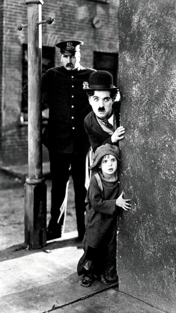 Charlie Chaplin and a child peeking from behind a wall with a police officer standing nearby