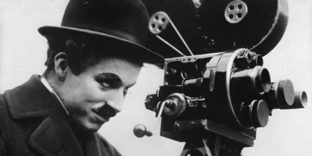 Charlie Chaplin wearing a bowler hat operating a vintage movie camera with focus and determination