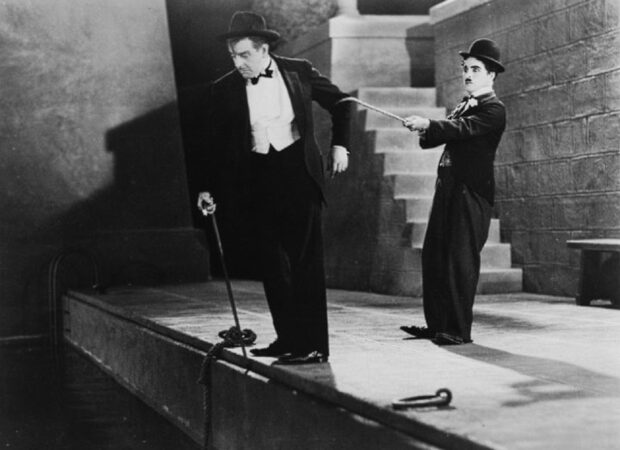 Charlie Chaplin character pulling a man with a cane on the dock in a classic silent film scene