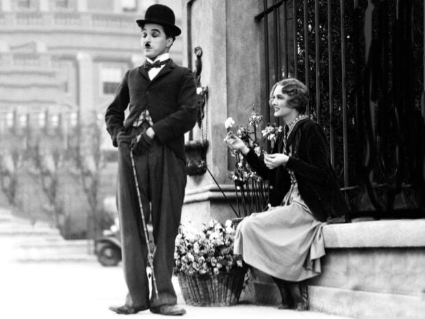 Charlie Chaplin is standing with a cane while a woman is sitting and holding flowers near a fence