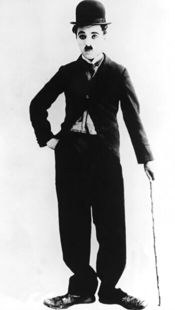 Charlie Chaplin stands with a cane wearing his iconic outfit and bowler hat in black and white image
