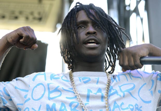 Chief Keef wearing a gold chain and light blue shirt performing with raised fists on stage