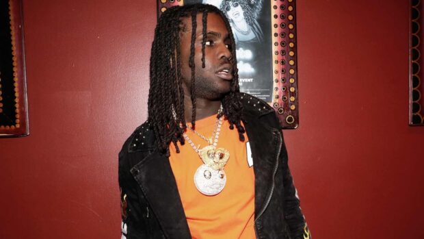 Chief Keef wearing a black jacket and orange shirt with chains against a red wall