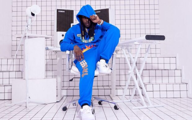 Chief Keef wearing a blue hoodie and pants sitting in a white tiled room