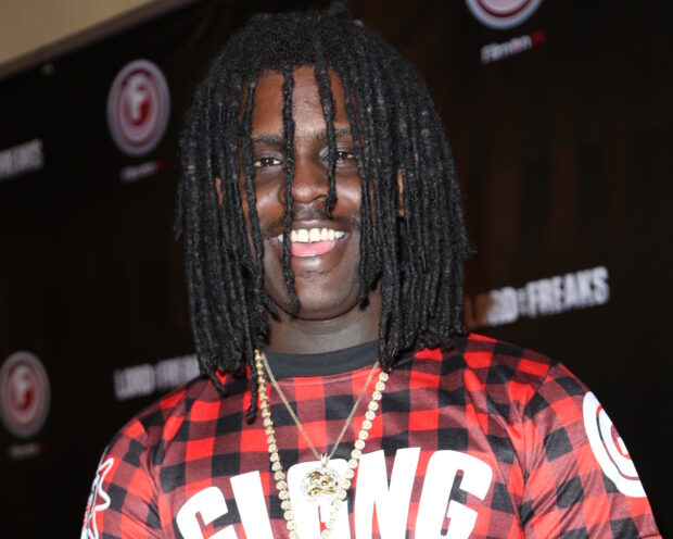 Chief Keef wearing a red checkered shirt and gold chain smiling at an event