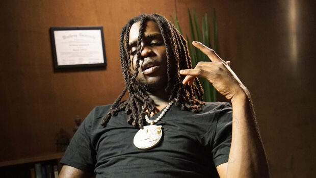 Chief Keef with dreadlocks and a large pendant necklace sitting indoors