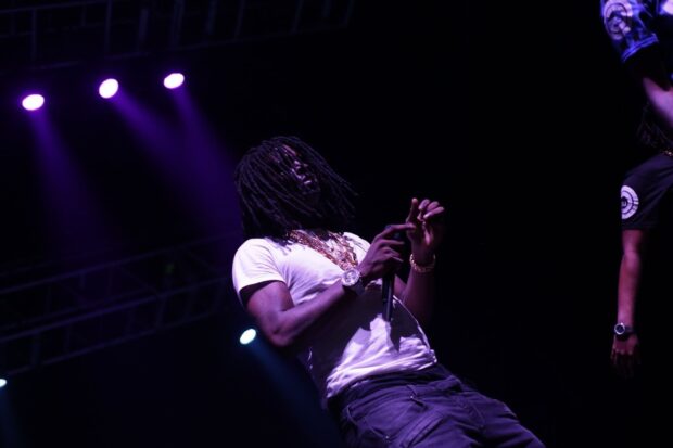 Chief Keef performing on stage wearing a white shirt and gold chains under purple lights
