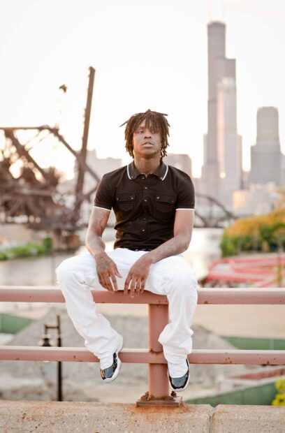 Chief Keef wearing a black shirt and white pants sitting on a railing near a city river