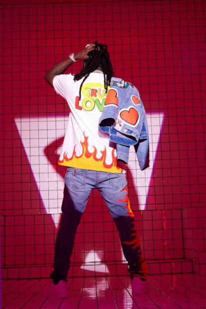 Chief Keef wearing flame jeans and a colorful shirt standing against a red tiled wall with a denim jacket on his shoulder