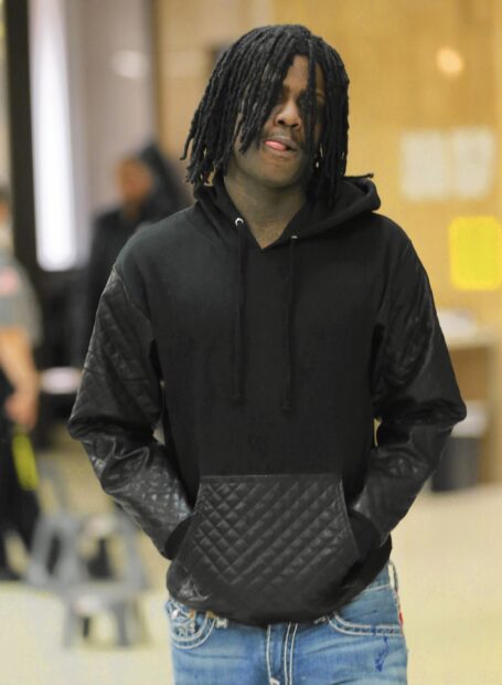 Chief Keef wearing a black hoodie with quilted leather patches and blue jeans standing indoors with hands in pockets