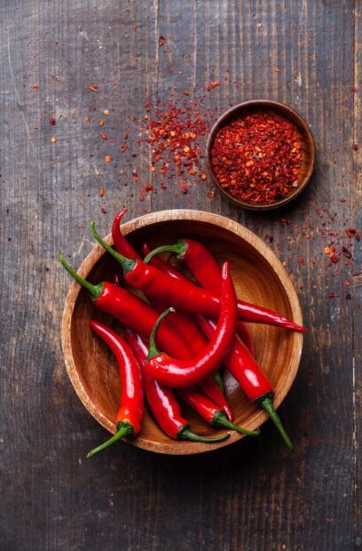 Fresh chili peppers in a wooden bowl with chili flakes on a rustic surface