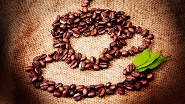 Coffee beans arranged to form a coffee cup shape with green leaves on a burlap surface