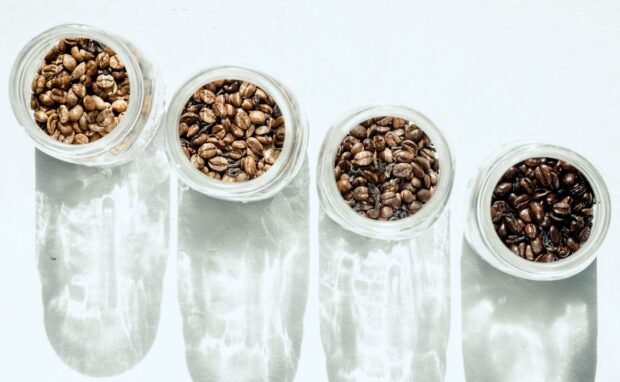 Different roasted coffee beans in glass jars casting shadows on white surface