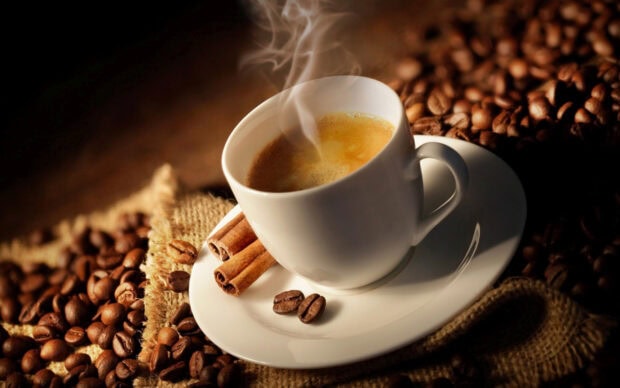 A steaming cup of coffee surrounded by coffee beans and cinnamon sticks on a rustic cloth