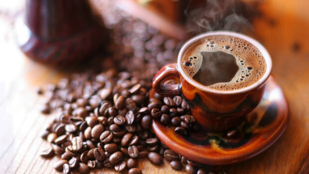A steaming cup of coffee surrounded by roasted coffee beans on a wooden surface