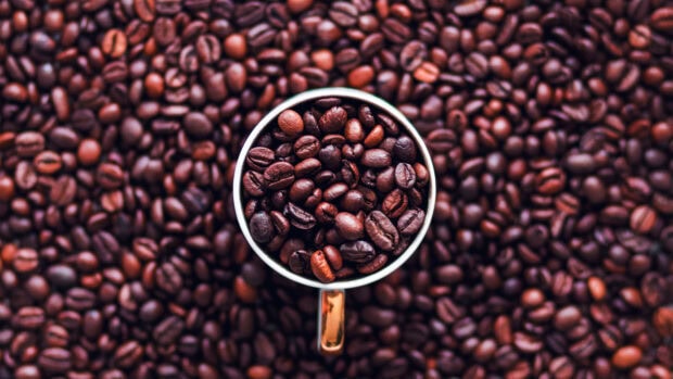 Coffee beans filling a cup surrounded by more coffee beans on a flat surface