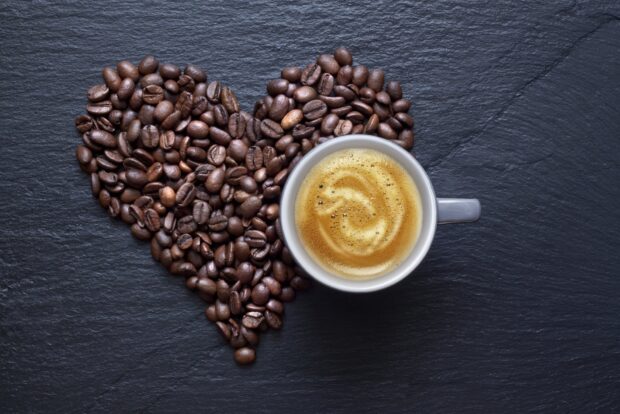 Heart shape made of coffee beans next to a cup of creamy coffee on a dark surface