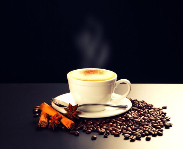 A steaming cup of coffee surrounded by coffee beans and cinnamon sticks on a black surface