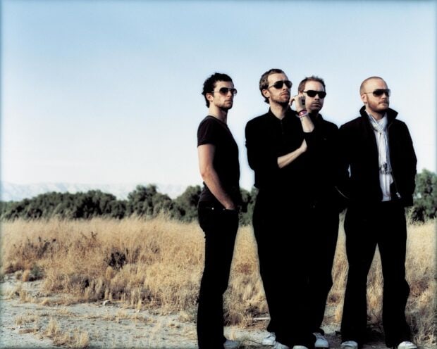 Coldplay band members standing outdoors in a dry field under clear sky