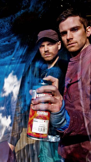 Two men holding a spray can with Coldplay in a reflective blue background