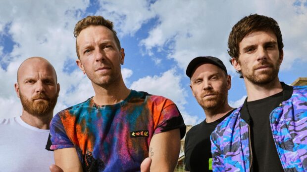 Coldplay band members posing outdoors with colorful clothes under a blue sky