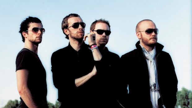 Coldplay band members standing outdoors in sunglasses and dark clothing on a clear day