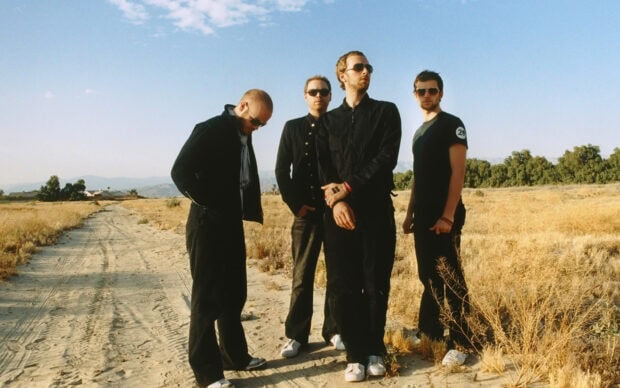 Coldplay members standing on a dirt road in a dry field with a clear blue sky in the background