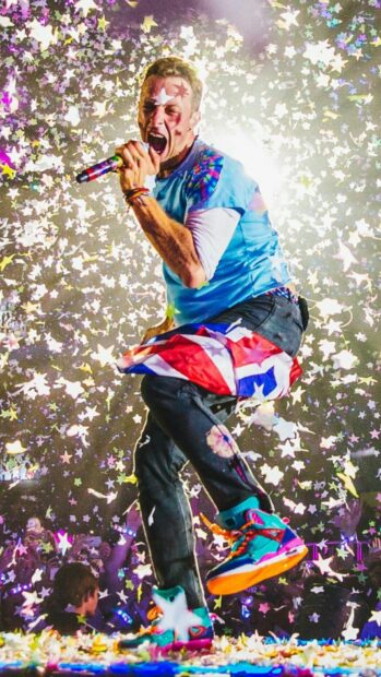 Coldplay performing energetically on stage with a flag and colorful confetti surrounding the singer
