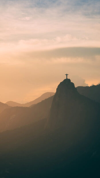 Corcovado Mountain is seen at sunset with the iconic statue on top surrounded by soft clouds