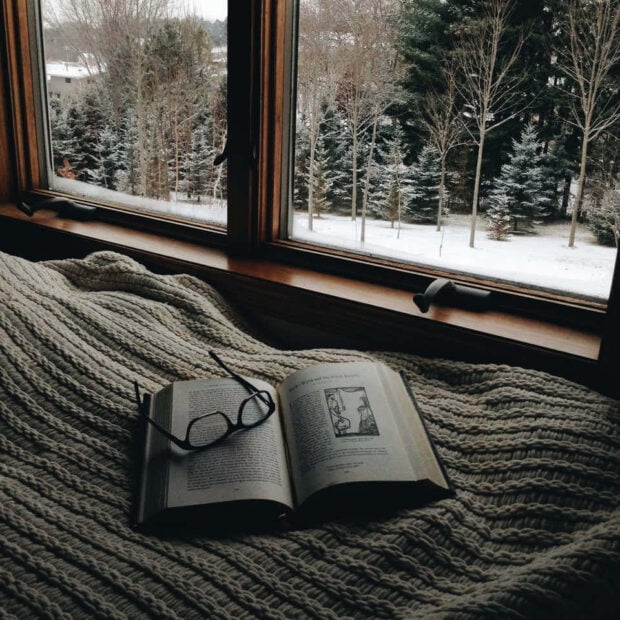 A cozy winter scene with a knitted blanket and open book near a snowy forest view through the window