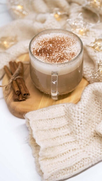 Hot cinnamon latte with cozy knitted sweater and cinnamon sticks on wooden board