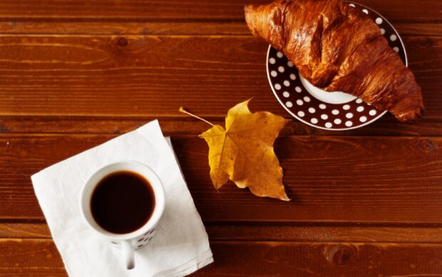 A croissant on a polka dot plate next to a cup of coffee and an autumn leaf on a wooden table