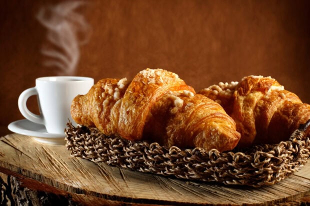A croissant on a woven basket next to a steaming cup of coffee on a wooden table