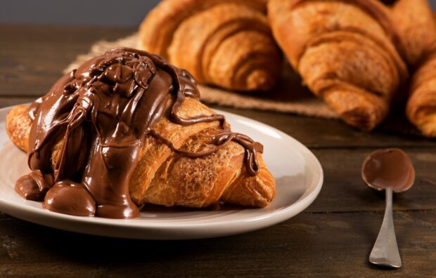 A croissant topped with melted chocolate spread on a white plate on a wooden table
