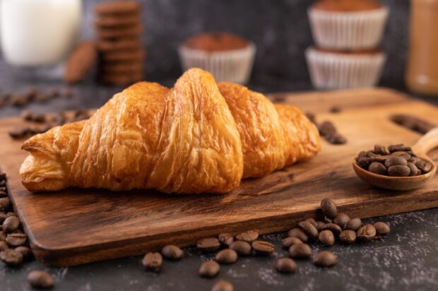 A fresh croissant on a wooden board surrounded by coffee beans and muffins in the background