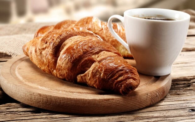 A fresh croissant placed on a wooden board next to a cup of coffee on a rustic table