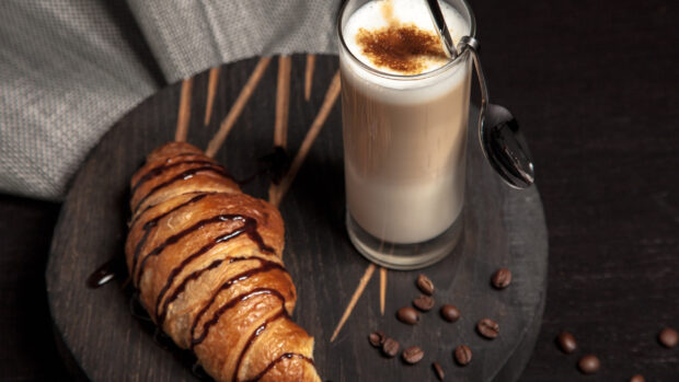 A croissant drizzled with chocolate sauce served on a wooden plate with coffee beans and a glass of frothy coffee