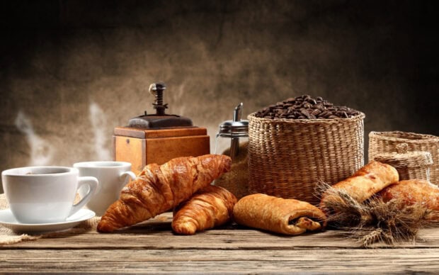 Fresh croissant and coffee beans placed on a wooden table with a vintage coffee grinder and steaming cups