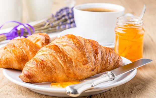 Fresh croissant with orange marmalade spread on a plate next to a cup of coffee and a knife