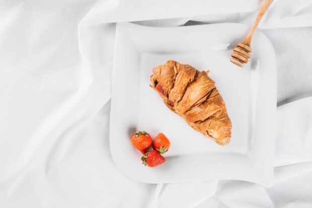A croissant with strawberries on a white plate with a honey dipper on a white cloth background